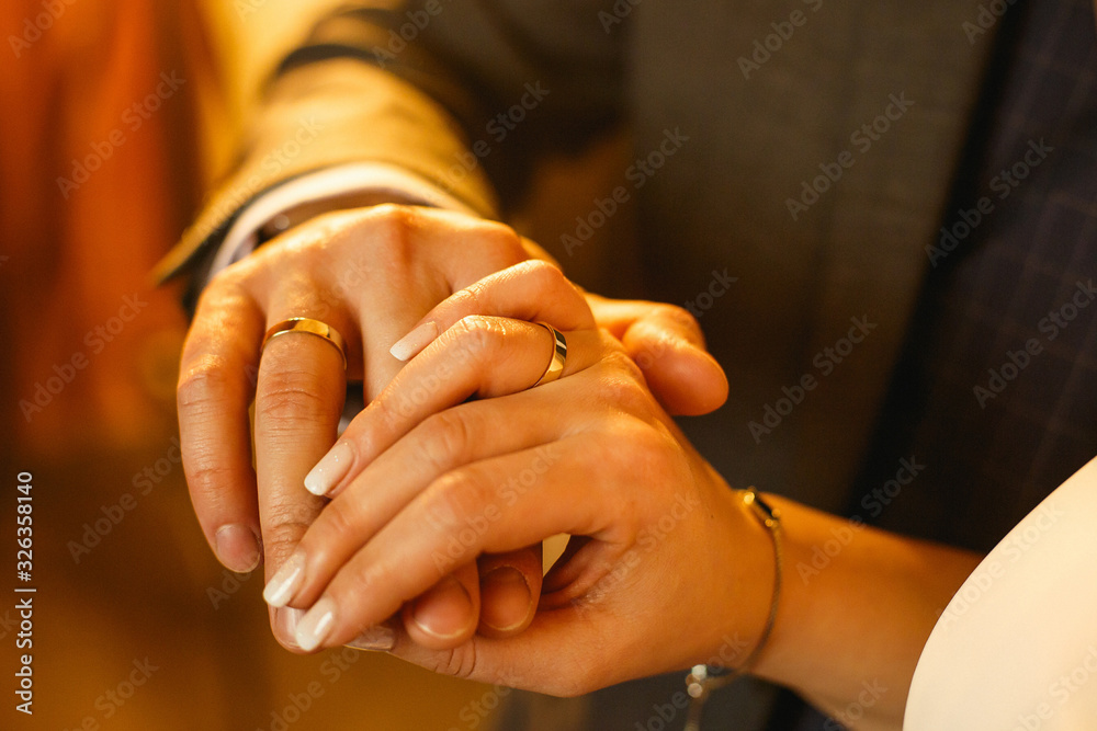 Fototapeta premium Two newlyweds hands, one female and one male, holding each other. On both hands a golden wedding ring. Male hand above. Selective focus. Close up. Warm golden light.