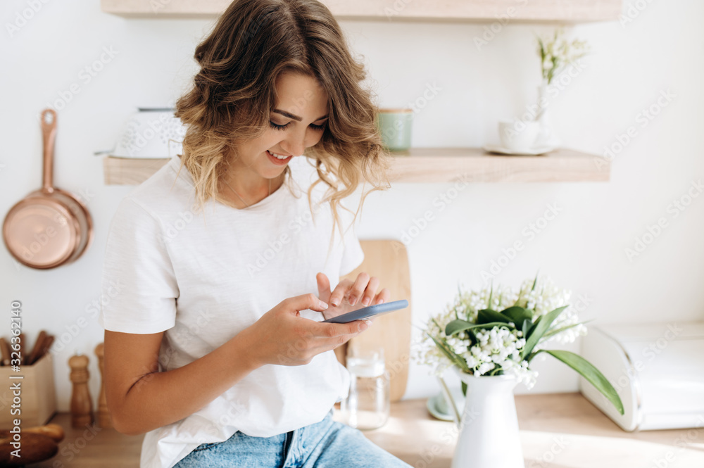 Smiling young girl is typing on her phone while sitting on a table in the kitchen