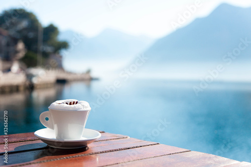 Fototapeta Naklejka Na Ścianę i Meble -  Cup of coffee on wooden table at sea beach. Authentic countryside cafe in rustic style. Morning breakfast with amazing view of winter lake and blue mountains. Cozy sunny still life. Copy space.