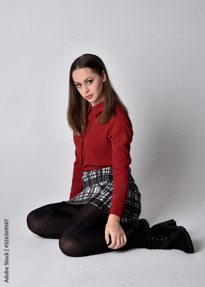 full length portrait of a pretty brunette girl wearing a red shirt and  plaid skirt with leggings and boots. Seated pose on a studio background.  Stock Photo