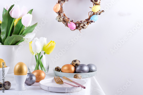 Holiday congratulation composition of served table with handmade painted eggs, baked cookies, fresh spring tulips flowers and festive wreath on a light grey wall background. Happy Easter concept.