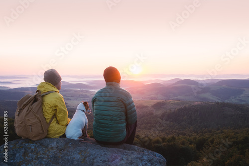 Two tourists sitting on the edge of the cliff with white dog against the backdrop of an incredible sunrise mountains. Landscape photography