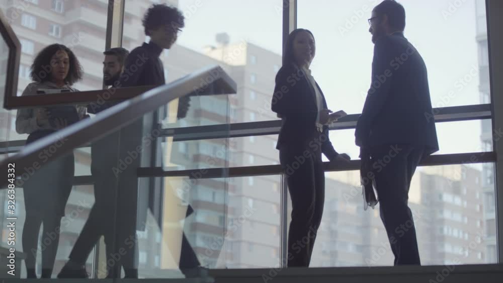 Slowmo of businesspeople standing on landing of staircase of office building with panoramic window and discussing work. Businesswoman with coffee in to-go cup walking down stairs Slowmo of businesspeople standing on landing of staircase of office building with panoramic window and discussing work. Businesswoman with coffee in to-go cup walking down stairs