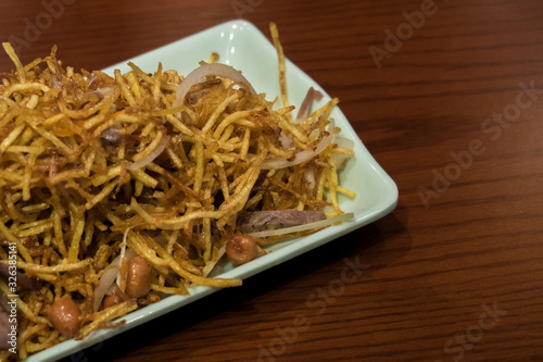 Chinese salad of fried thin chips of potatoes, onions and peanuts on a white plate on a brown wood table.