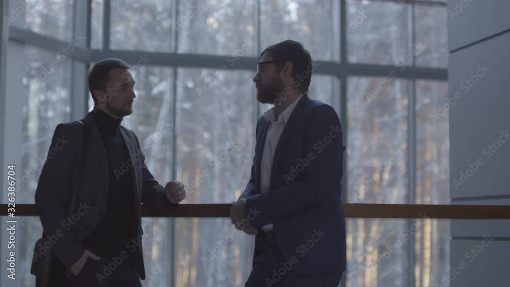 Slowmo shot of stylish businessmen with beards leaning on railing on staircase landing of office building with panoramic window and having conversation
