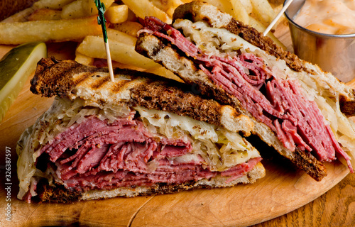 Reuben Sandwich, classic traditional American bar/pub menu item, on grilled rye bread, corned beef, Swiss cheese, sauerkraut and topped with thousand island dressing and french fries in background.