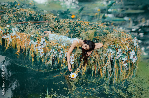 River Nymph in white vintage dress lies in boat decorated flowers yellow willow branches, enjoy silence relax. long flowing dark hair. touches hand lily. Backdrop autumn orange nature green water lake