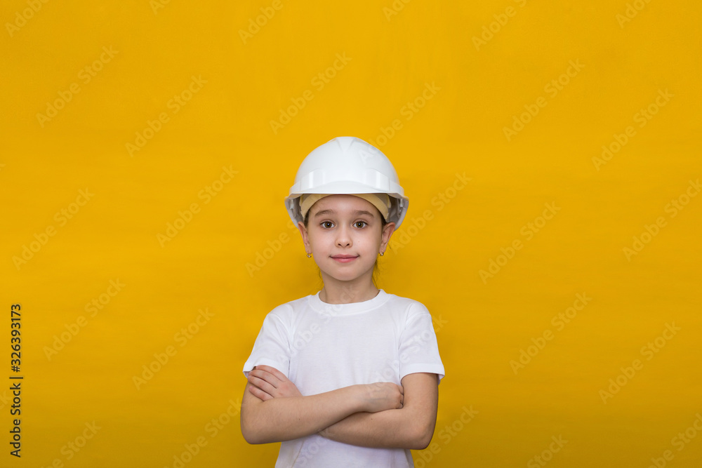 smiling little girl in a construction white helmet crossed her arms over her chest on a yellow background