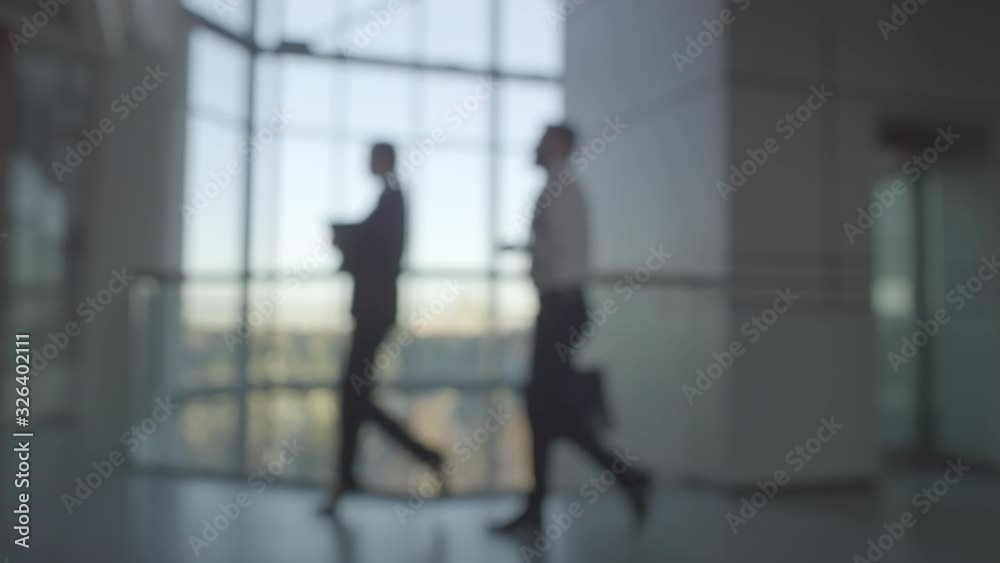 Blurry shot of unrecognizable businesswoman in suit and businessman with briefcase walking along hallway of modern office building. Businessman on scooter passing by camera Blurry shot of unrecognizable businesswoman in suit and businessman with briefcase walking along hallway of modern office building. Businessman on scooter passing by camera