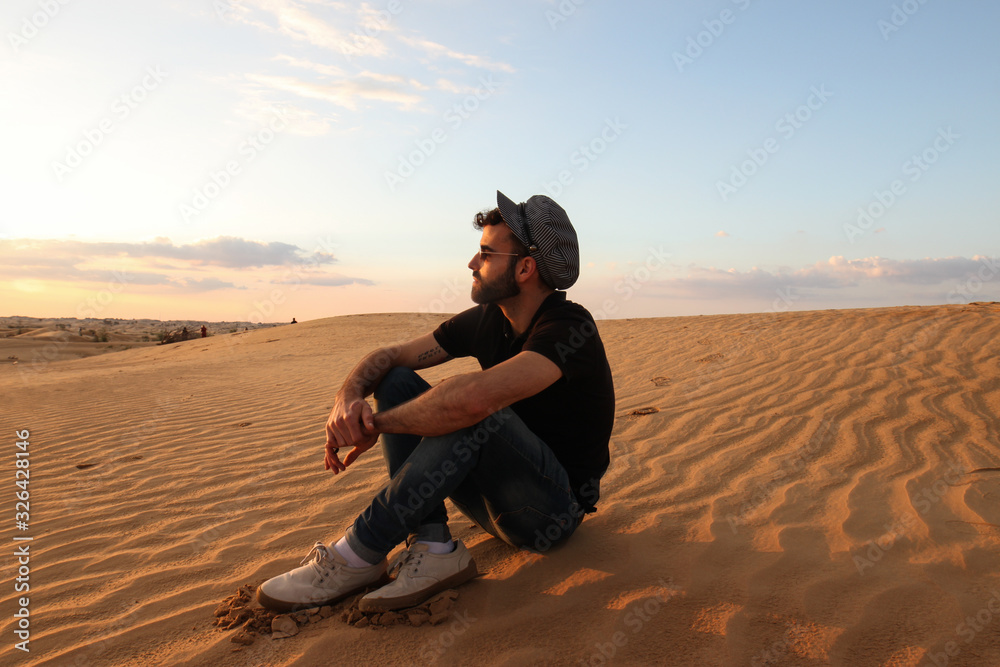Young hipster man with hat and sunglasses sitting on desert dunes during sunset