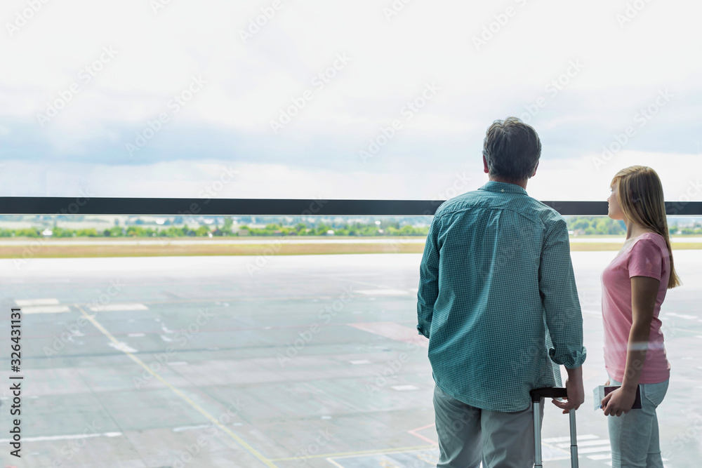 Fototapeta premium Rear view of mature man looking through the window with his daughter in airport