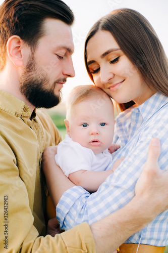 portrait of a happy young family, where mom and dad hug each other, holding their little child between them in the rays of the sunset, summer sun