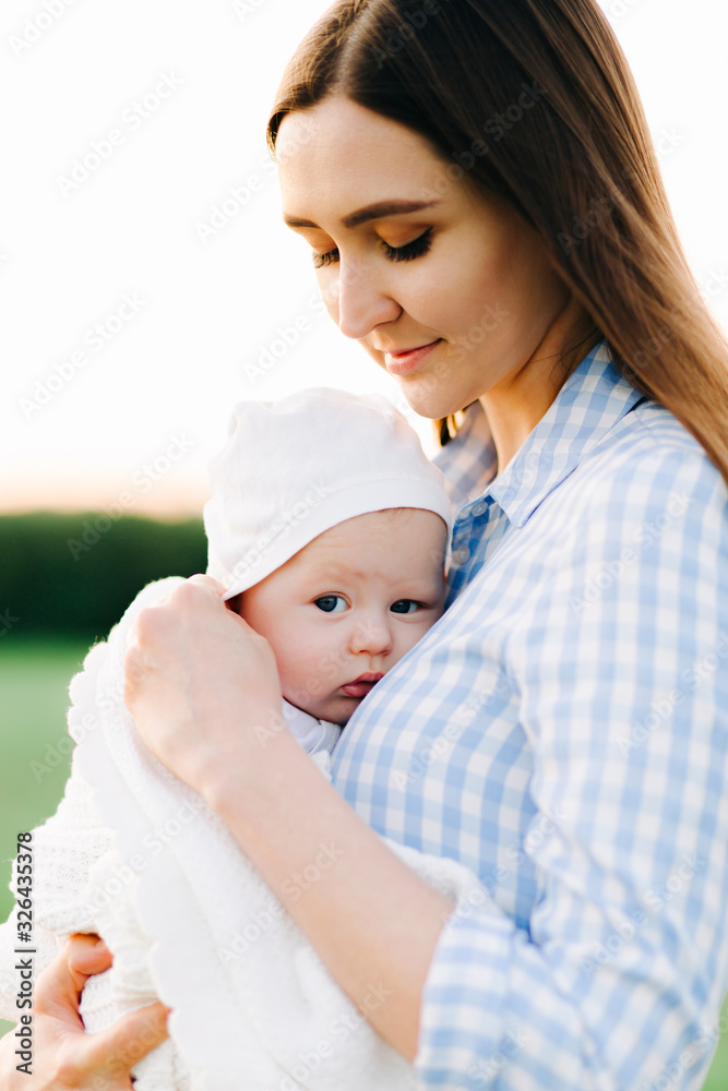 portrait of a young mother in a blue checked dress, holding a small child to her chest, in the rays of the setting summer sun