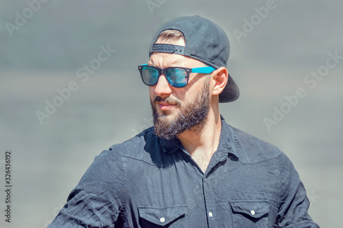 portrait of a stylish young man with a beard in a baseball cap, sunglasses and a black shirt on a blurry gray background on a sunny summer day