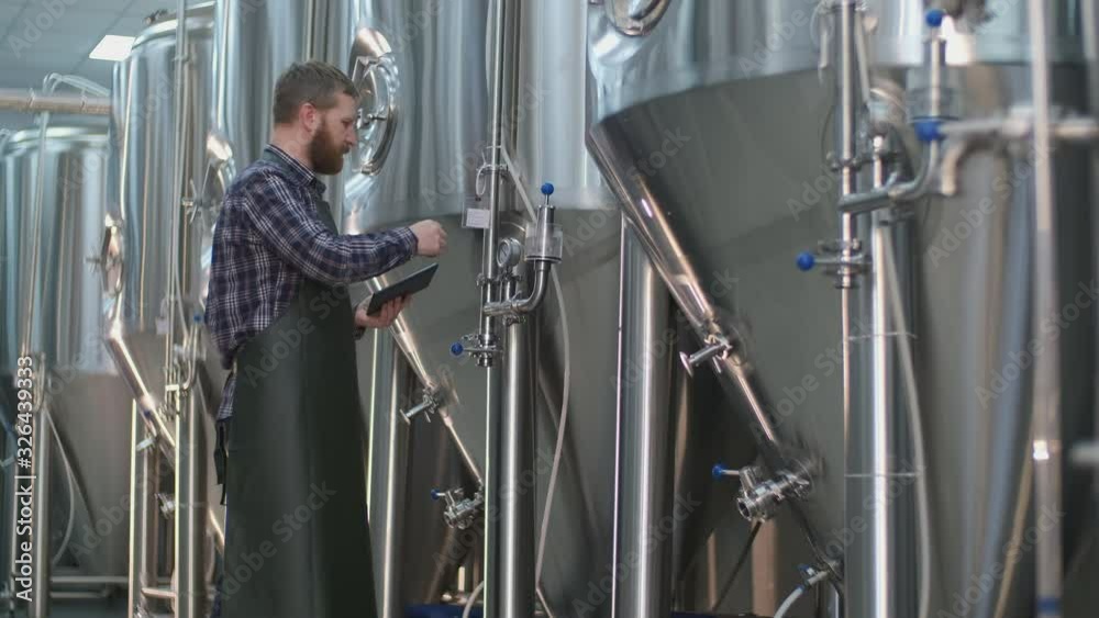 Brutal Male brewer with a beard controls the readings of devices on beer tanks using a tablet. Craft beer production