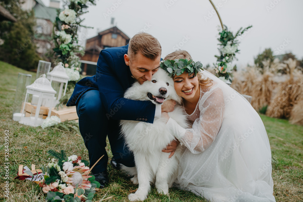 Newlyweds play with dog on background of lawn and house.