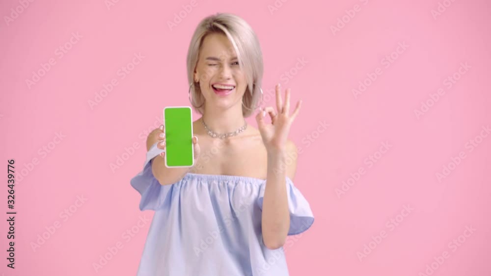 happy girl pointing with finger at smartphone and showing okay isolated on pink happy girl pointing with finger at smartphone and showing okay isolated on pink