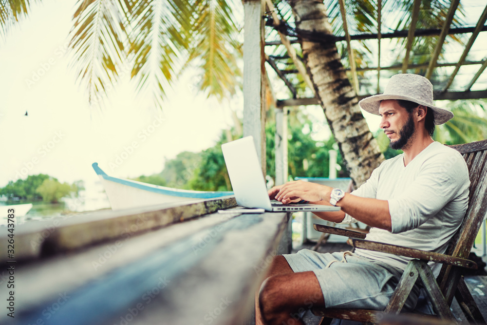 Technology and travel. Working outdoors. Freelance concept. Bearded young man using laptop in cafe on tropical beach. - obrazy, fototapety, plakaty 