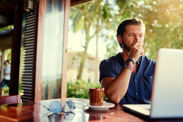 Freelance concept. Young  bearded man using laptop while sitting on summer terrace.