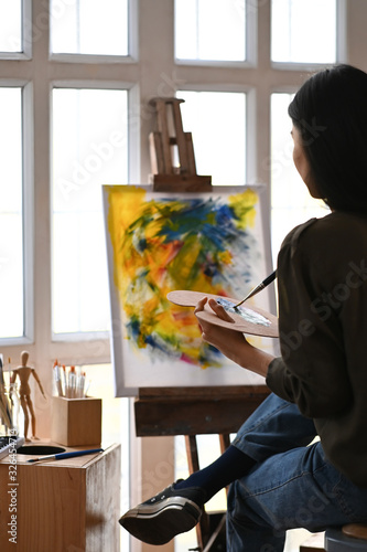 Young artist girl sitting in front the drawing canvas while painting an oil color at the modern studio. Beautiful artist and gallery studio concept.