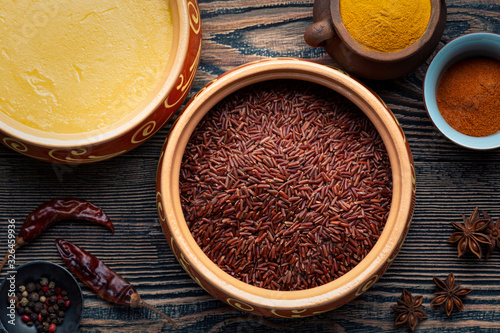Ghee or clarified butter in ceramic bowls and red rice with different spices on an old wooden table.