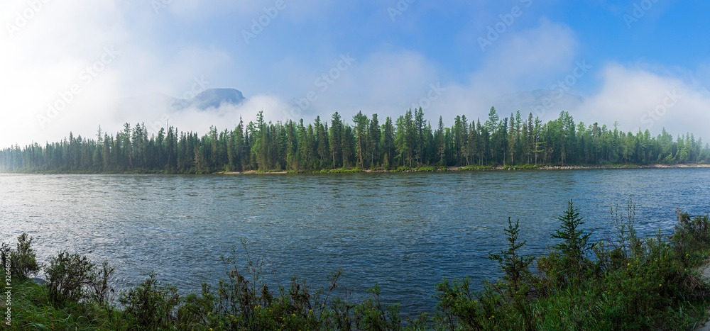 Panorama of the shore of a mountain river. Mountains are partially hidden by rising morning fog.