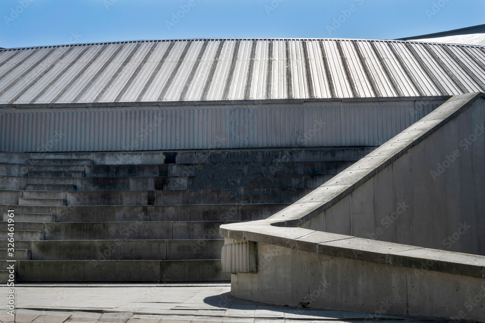 Edificio gris construido con cemento y con techo de aluminio formando en conjunto efectos lineales.