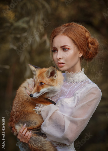 Close up portrait of amazing cute young woman in beautiful fairy-tale image with fire red hair in white lace dress looks far away and holds small red and white fox in beautiful yellow autumn forest