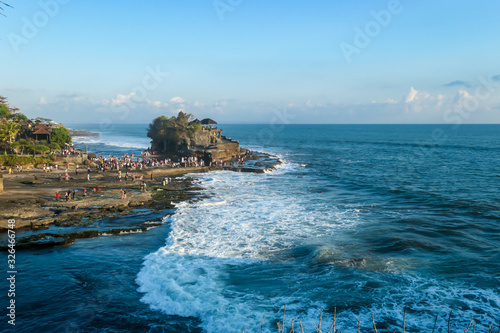 Crowds visiting the Tanah Lot Temple, Bali, Indonesia, located on the cliffs by the seashore. The waves are splashing on the cliffs and smaller rocks. Clear and sunny day. Power of the nature