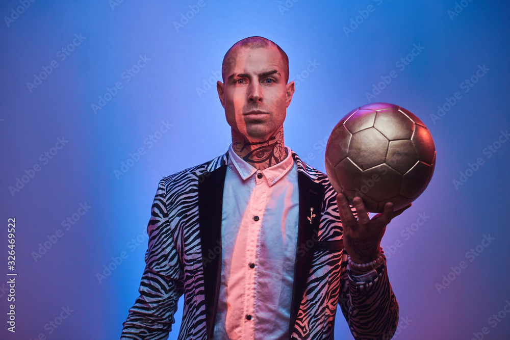 Plakat Elegant, handsome, tattooed, bald male model posing in a studio for the photoshoot wearing fashionable custom made zebra striped style tuxedo and rose patterned shirt, holding a golden soccer ball in