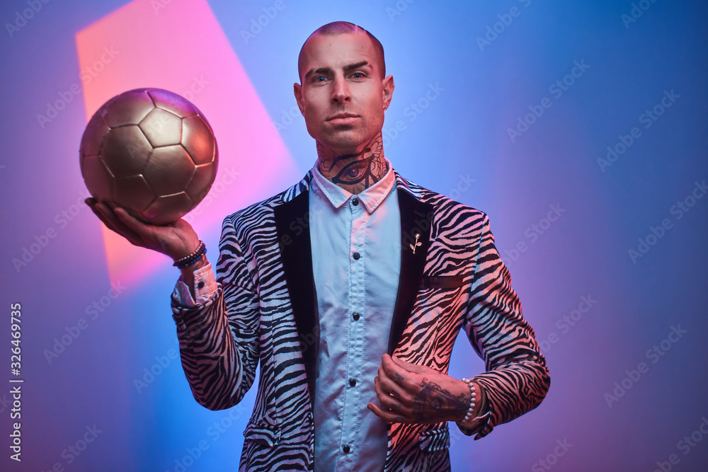 Plakat Fashionable, handsome, tattooed, bald male model posing in a studio for the photoshoot wearing fashionable custom made zebra striped style tuxedo and rose patterned shirt, looking on a golden soccer