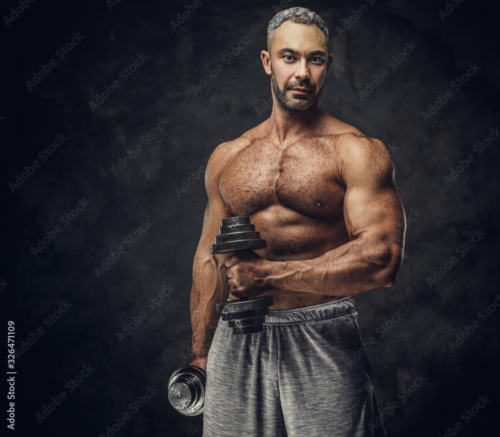 Powerful, adult, fit muscular caucasian man coach posing for a photoshoot in a dark studio under the spotlight wearing grey sportswear, showing his muscles and putting up a dumbbells looking confident