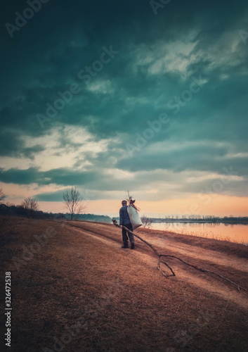 Rural evening scene, vertical photo with a lone nomad man carrying firewood and a bagful on his shoulder, going home walking a country dirt road near the lake. Moody and dramatic view.