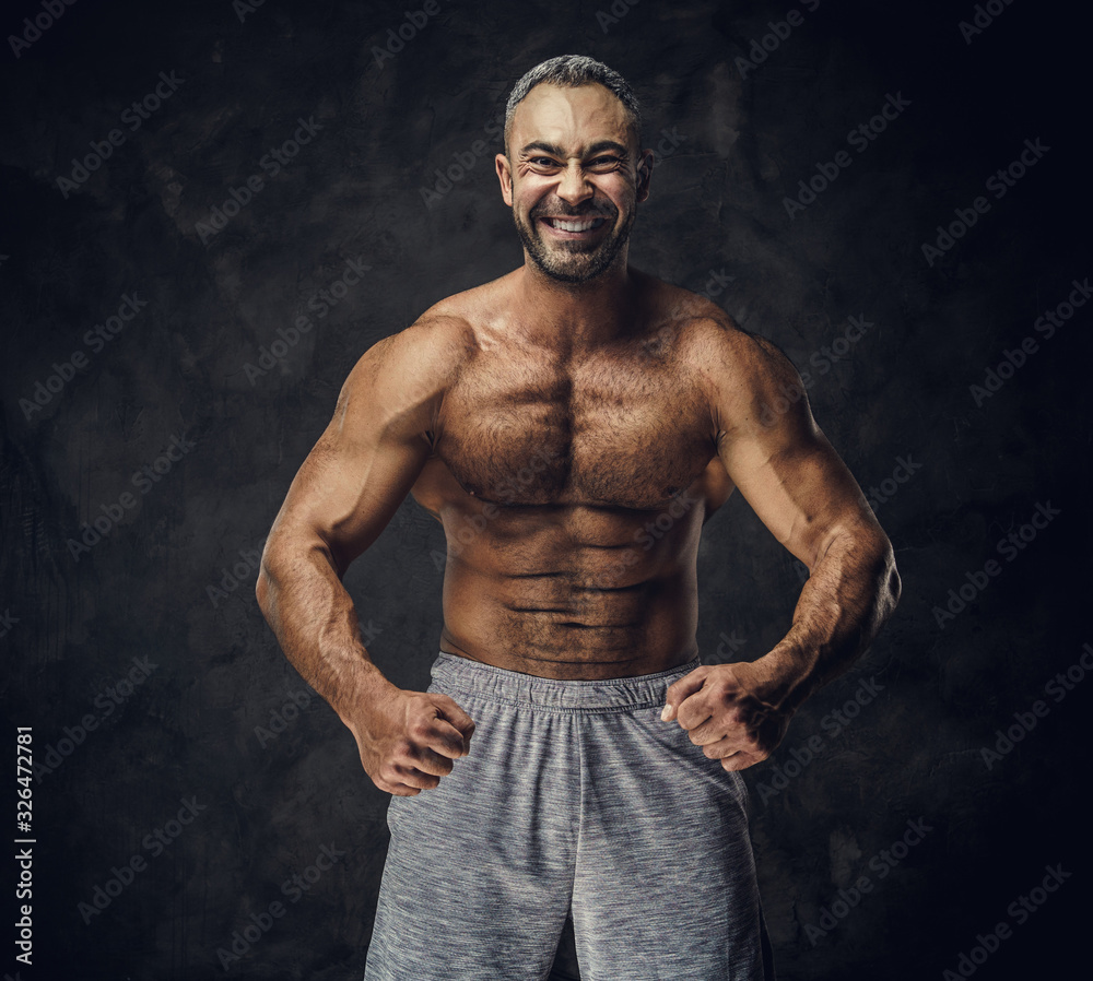 Strong, adult, fit muscular caucasian man coach posing for a photoshoot without his shirt in a dark studio under the spotlight wearing sporty shorts, showing his muscles looking powerful and smiling Strong, adult, fit muscular caucasian man coach posing for a photoshoot without his shirt in a dark studio under the spotlight wearing sporty shorts, showing his muscles looking powerful and smiling