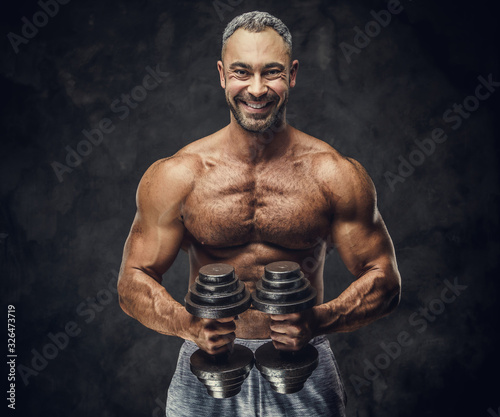 Heavyweight, adult, fit muscular caucasian man coach posing for a photoshoot in a dark studio under the spotlight wearing grey sportswear, showing his muscles and putting up a dumbbells looking
