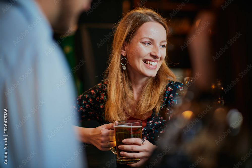Portrait of cheerful caucasian blonde standing in pub, holding pint of beer and chatting with friends. Nightlife.