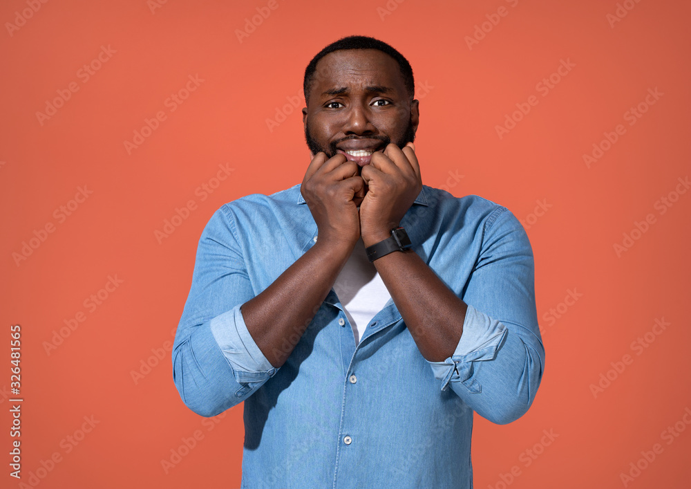 Worried man bites nails, looks nervous. Photo of african man in casual outfit on coral background. Worried man bites nails, looks nervous. Photo of african man in casual outfit on coral background.