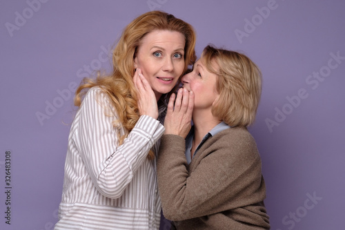 Two beautiful caucasian mature women telling secret on a colored background.