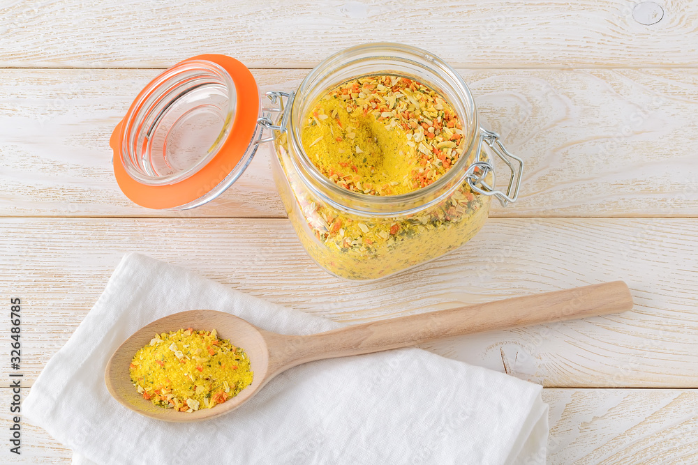 Universal seasoning in a clip top glass jar and in a large wooden spoon on a white wooden kitchen table. Seasoning with salt and dry vegetables. Cook at home. Universal seasoning in a clip top glass jar and in a large wooden spoon on a white wooden kitchen table. Seasoning with salt and dry vegetables. Cook at home.