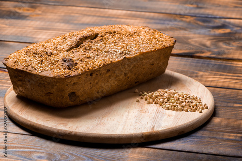 A loaf of freshly baked buckwheat bread lies on a round kitchen board on a brown wooden table. A handful of green buckwheat seeds are scattered nearby. Harmless baking for vegans and vegetarians