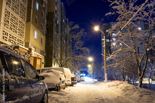 Snow-covered city courtyard in the late evening. Magical landscape. Snow-covered cars parked along high-rise buildings, lights of lanterns. Winters Tale in Donetsk, Ukraine. Scenic view from road