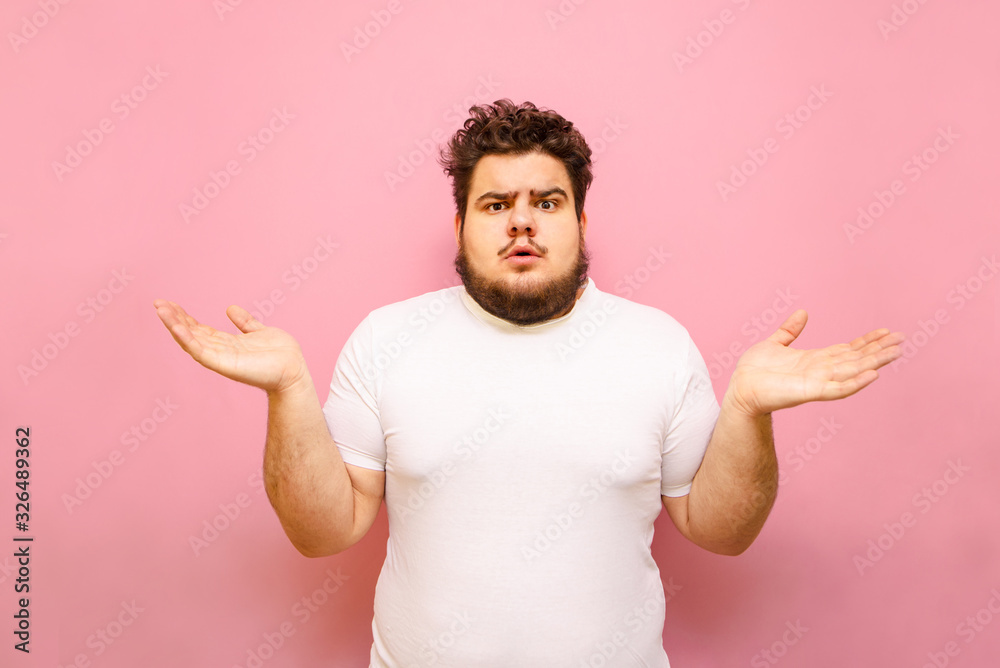 Puzzled young overweight man stands on a pink background and spreads his arms out to the sides. Confused fat guy does not know, surprise looking at the camera, isolated. Copy space Puzzled young overweight man stands on a pink background and spreads his arms out to the sides. Confused fat guy does not know, surprise looking at the camera, isolated. Copy space