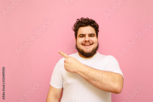Happy young man with overweight and beard isolated on a pink background, looks into the camera and smiles, points his finger away at copy space. Fat guy in a white shirt shows an empty space
