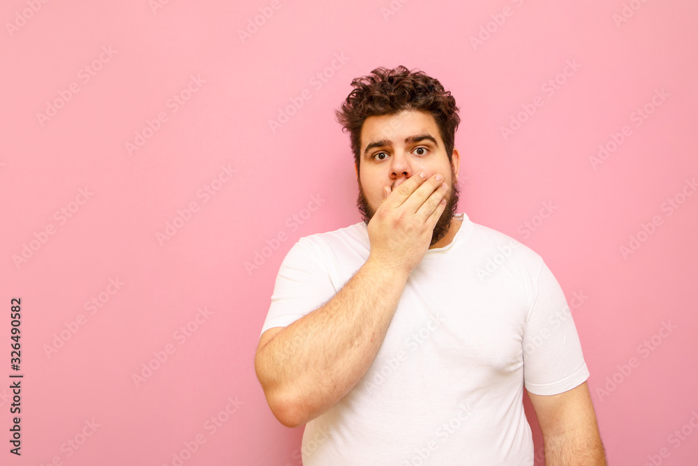 Surprised overweight guy stands on a pink background, closes his mouth in astonishment and looks into the camera. Fat funny curly young man with shocked face covering his mouth. Surprised overweight guy stands on a pink background, closes his mouth in astonishment and looks into the camera. Fat funny curly young man with shocked face covering his mouth.