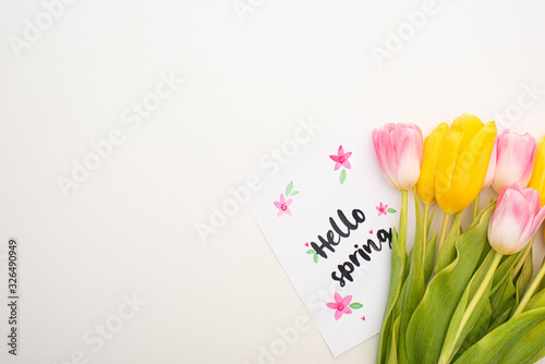 Top view of card with hello spring lettering near yellow and pink tulips on white background
