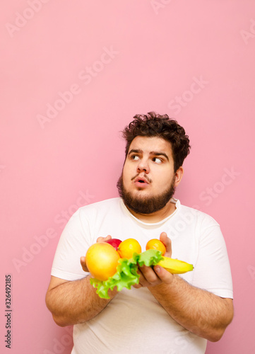 Surprised fat man on diet standing with fruits in hands on pink background and looking up at copy space with shocked face.Overweight guy looks on empty space and holds fruits and vegetables in hand