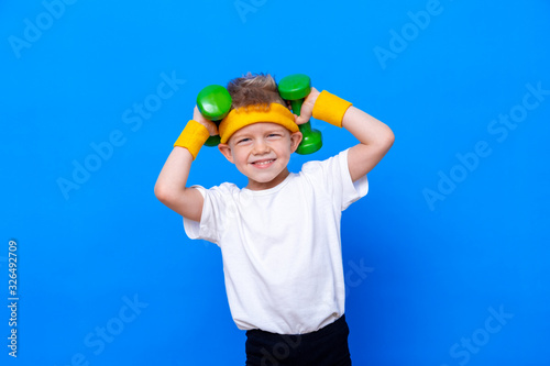 Fitness child. Portrait of sporty little boy with dumbbells over blue studio background. Gym workout. Child sportsman. Childhood activity. Sport. Fitness, health and energy. Success