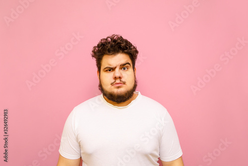 Portrait of a funny fat man in a white t-shirt on a pink background, looking into the camera with a serious funny face. Big guy isolated on pink background. Copy space
