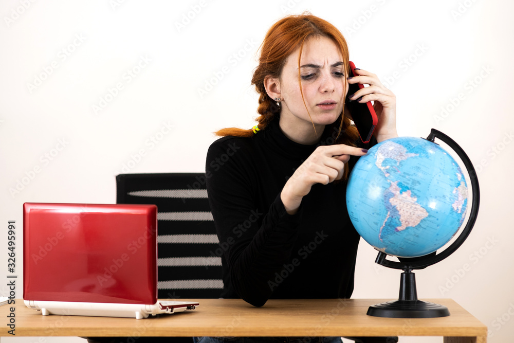 Young travel agent woman sitting behind working desk with laptop computer and geographic globe of the world talking on a cell phone. Young travel agent woman sitting behind working desk with laptop computer and geographic globe of the world talking on a cell phone.