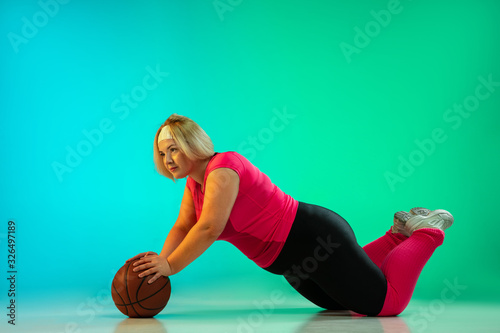 Young caucasian plus size female model's training on gradient green background in neon light. Doing workout exercises with ball, basketball. Concept of sport, healthy lifestyle, body positive